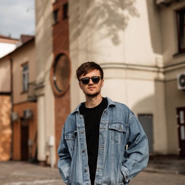 Güneşli bir yaz gününde, klasik binaların yakınında mavi bir ceketle şık güneş gözlüklü şık bir genç adam modeli. Çekici adam şehirde gezip duruyor. Moda erkek kıyafetleri.