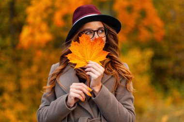 Şık gözlüklü, zarif bir gülümsemesi olan, uzun montlu zarif bir şapkası olan oldukça mutlu genç bir kadın yüzünün yanında turuncu bir akçaağaç yaprağı tutuyor. Parkta sonbahar yaprağıyla poz veren mutlu hippi kız..