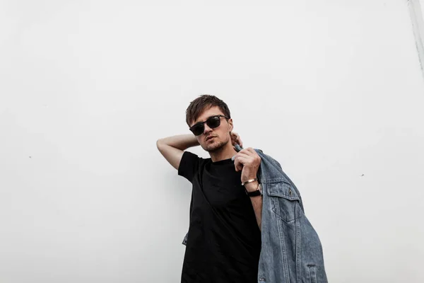 Young urban american man hipster with a beard with a hairstyle in a stylish denim blue jacket in a black t-shirt in fashionable sunglasses posing near a vintage white wall in the city. Handsome guy. — Stock Photo, Image