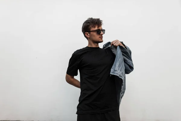 Modische junge Mann Hipster zieht eine stylische Jeansjacke an. attraktiver Typ in trendiger Kleidung mit Sonnenbrille, der auf einer Straße in der Stadt in der Nähe einer weißen Mauer posiert. Jugend neue Kollektion von Herrenbekleidung. — Stockfoto