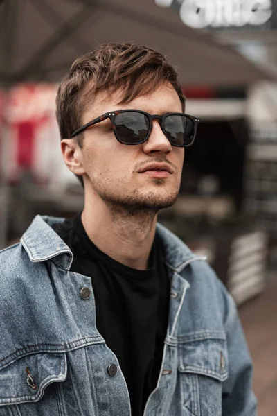 Portret amerykańskiego przystojnego młodzieńca z brodą w modnych okularach przeciwsłonecznych w stylowej niebieskiej kurtce jeansowej w t-shircie na ulicy w wiosenny dzień. Atrakcyjny hipsterski facet spaceruje po mieście. — Zdjęcie stockowe