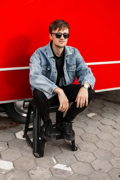 ビンテージサングラスの流行の黒いパンツでファッショナブルなデニムジャケットのクールなハンサムな若い男は、市内の近代的な赤いバンの近くのはしごに座って休んでいます。スタイリッシュな魅力的な男ヒップスター屋外 — ストック写真