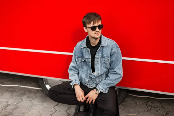 Modern urban young hipster man in fashionable denim clothes in trendy black sunglasses sits near a vintage red van on the street on a summer day. American guy model resting in the city. — Stock Photo, Image