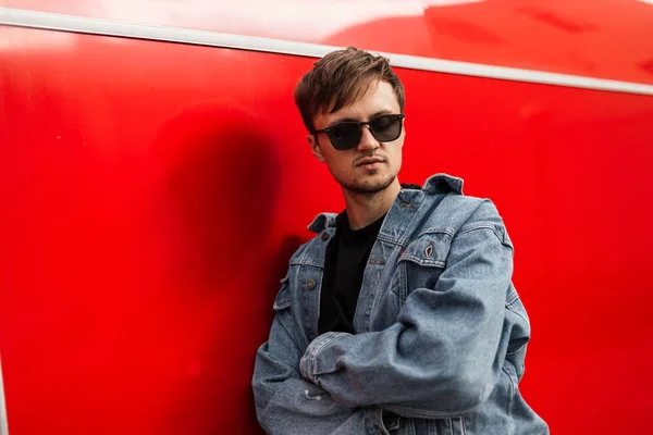 El joven hipster cool de moda con una chaqueta de mezclilla en una camiseta negra con gafas de sol con un peinado posa cerca de una pared roja de metal vintage. Un tipo con estilo americano descansando en la ciudad. Estilo de calle . — Foto de Stock