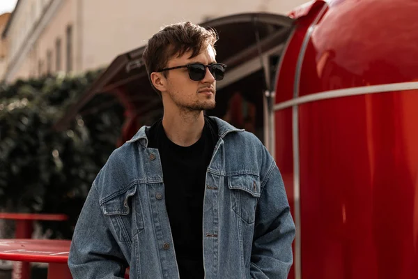 Bonito hombre hipster joven con estilo en una chaqueta de mezclilla en una camiseta negra en gafas de sol con un peinado se encuentra cerca de una camioneta roja de metal vintage. La modelo urbana de moda está descansando en la ciudad. Moda callejera . — Foto de Stock