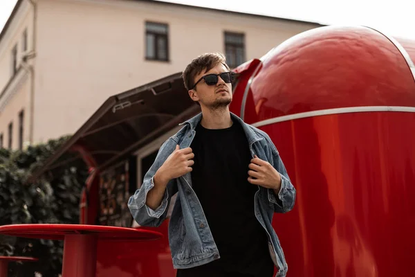 El joven hipster urbano se quita una chaqueta de mezclilla de moda. Atractiva modelo de chico en gafas de sol posa en el fondo de una furgoneta roja con comida en la ciudad en un soleado día de primavera. Moda masculina de moda . — Foto de Stock