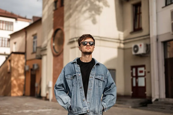 Elegante hombre hipster joven atractivo en una chaqueta de mezclilla azul de moda en gafas de sol negras disfruta de un paseo por una calle de la ciudad en un día soleado de primavera. Guapo chico modelo camina cerca de edificios . — Foto de Stock