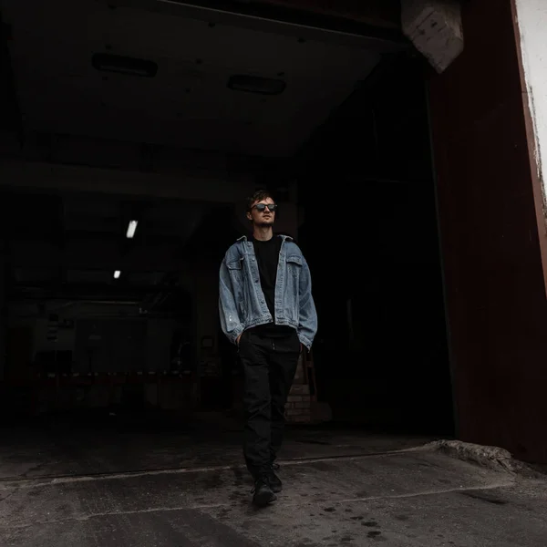 Fiatal amerikai menő férfi hipster divatos ifjúsági ruhák napszemüvegben jön ki egy sötét raktárból. Jóképű városi fickó sétálgat a városban. Utcanév. — Stock Fotó