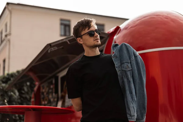 Joven de moda con un peinado elegante en una camiseta negra de moda en gafas de sol vintage con una chaqueta de mezclilla posando cerca de una moderna furgoneta de comida roja en la calle. chico urbano hipster paseos por la ciudad . — Foto de Stock