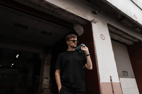 Jovem legal em óculos de sol elegantes em uma jaqueta jeans azul vintage em uma camiseta preta posa perto de um prédio na rua. Um tipo moderno. Coleção de primavera juvenil de roupas e acessórios masculinos — Fotografia de Stock