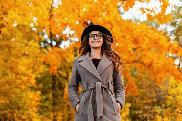 Mooie jonge vrouw met een positieve glimlach in een stijlvolle jas in een modieuze hoed met trendy bril staat in het park en geniet van het geel-oranje blad. Joyful hipster meisje wandelingen in het bos. — Stockfoto