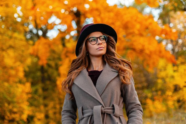 Modern cute young hipster woman with a stylish hairstyle in a vintage hat in fashionable glasses in an elegant coat walks on a the autumn park. Trendy girl model enjoys a walk through the woods. — Stock Photo, Image