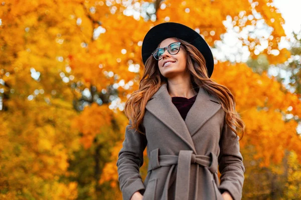 Mulher hipster jovem alegre em outerwear na moda em um chapéu elegante em óculos elegantes caminha no parque. Menina feliz com um sorriso bonito gosta do fim de semana. Coleção de outono de roupas femininas . — Fotografia de Stock