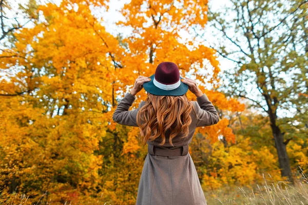 Вид спереду молодої жінки в стильному пальто в модному капелюсі в осінньому парку з деревами з жовто-оранжевим листям. Дівчина вирівнює розкішний капелюх у лісі. Осіння колекція жіночого верхнього одягу — стокове фото