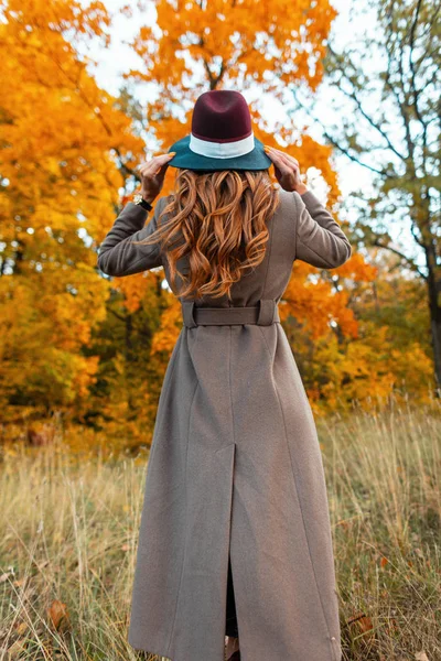 Achteraanzicht van een modieuze jonge vrouw in een stijlvolle lange jas met een elegante hoed met een trendy kapsel in een herfstpark tussen bomen met sinaasappelbladeren. Modern meisje loopt door het bos. — Stockfoto