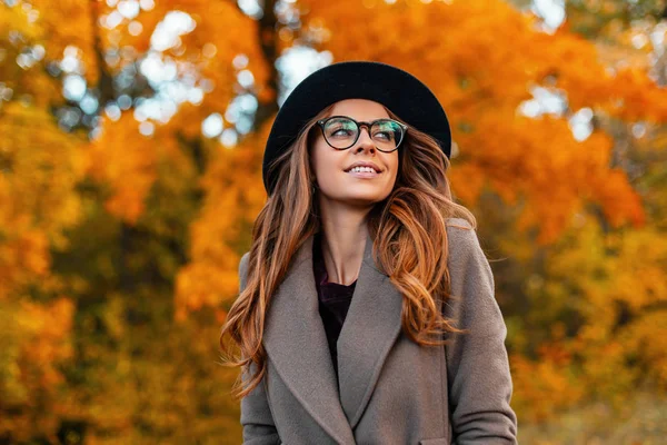 Досить молода елегантна жінка модель в розкішному капелюсі з окулярами в пальто з красивою посмішкою гуляє в парку серед дерева з апельсиновим листям. Сучасна весела дівчина хіпстер позує в лісі . — стокове фото
