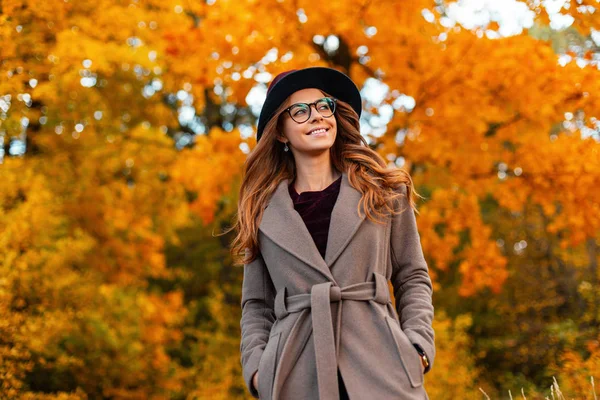 Mujer joven positiva con una hermosa sonrisa con el pelo rizado en un sombrero elegante en un abrigo elegante en gafas de moda disfruta del fin de semana en el parque de otoño. Linda chica hipster feliz es relaja al aire libre . — Foto de Stock