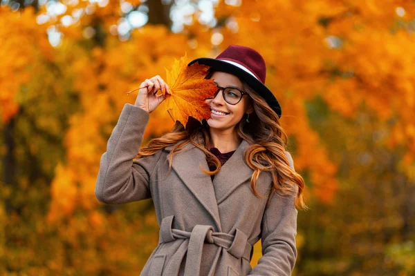 Joyeux jeune femme à la mode dans des lunettes dans un chapeau vintage dans une pose de manteau élégante avec une feuille d'automne orange-jaune à l'extérieur dans un parc. Joyeuse fille avec un sourire mignon tient une feuille d'érable dorée près du visage . — Photo