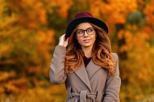 Jolie jeune femme hipster avec une coiffure élégante dans un chapeau vintage dans des lunettes à la mode dans un manteau élégant se promène sur un parc d'automne. attrayant fille modèle bénéficie d'une promenade dans les bois . — Photo