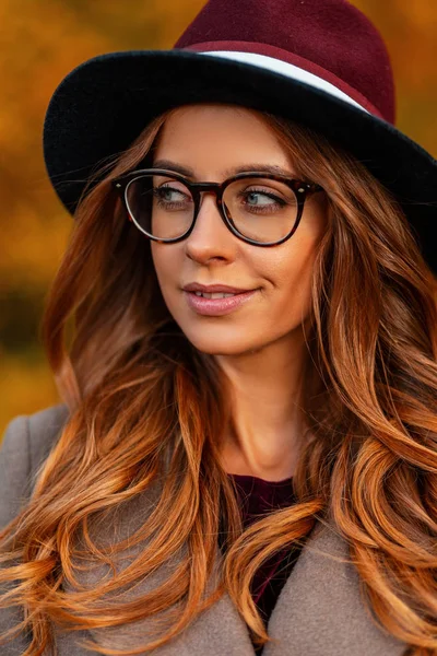 Retrato de una atractiva mujer hipster joven con el pelo castaño en un elegante sombrero elegante en un abrigo elegante en gafas vintage al aire libre en un parque. Moda hermosa chica modelo de moda. Estilo otoño . — Foto de Stock