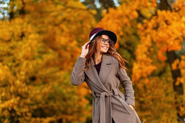Beautiful young woman model with a cute smile in a stylish coat in a fashionable hat in trendy glasses stands in the park and enjoys the yellow-orange foliage. Happy hipster girl posing in the forest. — Stock Photo, Image