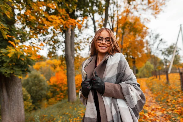 都会の外の公園でポーズをとる流行のコートでスタイリッシュなメガネでファッショナブルなニットスカーフで楽しい若いヒップスター女性。前向きな笑顔で陽気な女の子モデルが森の中を歩いています。秋. — ストック写真