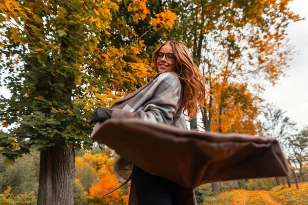 Ganska positiv ung kvinna i fashionabla kläder snurrar i höstskogen. Glada flicka modell i en stickad halsduk i en lång rock njuter av en promenad i parken bland träden med apelsinblad. — Stockfoto