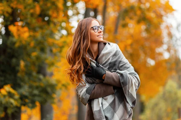 Mulher hipster europeia em um cachecol de malha na moda em óculos elegantes em um casaco na moda posando em um parque fora da cidade. Menina com um sorriso bonito caminha na floresta. Roupa de outono feminino elegante — Fotografia de Stock