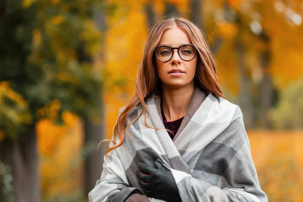 Porträtt av en ung attraktiv ung kvinna hipster i snygga glasögon i en rock i en vintage stickad halsduk på en bakgrund av gyllene lövverk i parken. Ganska vacker flicka njuter av en varm höstdag — Stockfoto