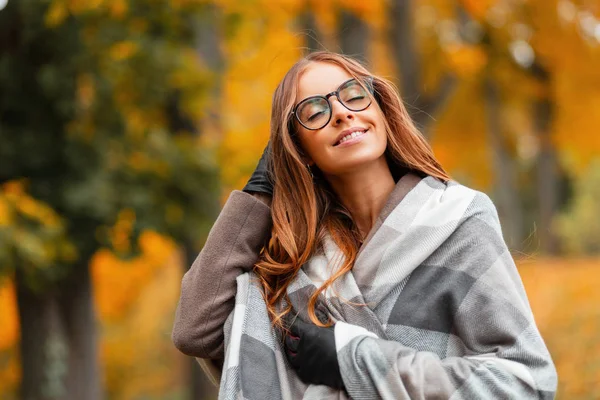 Портрет молодой веселой молодой женщины с милой улыбкой в стильных очках в пальто в винтажном вязаном шарфе на фоне золотой листвы в парке. Позитивная девушка наслаждается теплым осенним днем — стоковое фото