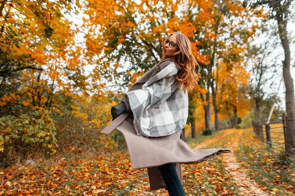 Elegant young woman in autumn stylish clothes is spinning in the park. Joyful girl model in a knitted scarf in a fashionable long coat enjoys a walk in the woods among the trees with orange leaves. — Stock Photo, Image