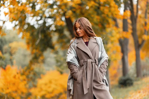 Stylish european young woman in a fashionable coat in a trendy checkered knitted scarf walks in the park with orange foliage. Attractive girl relaxes outdoors on a warm October day. Autumn clothes. — Stock Photo, Image