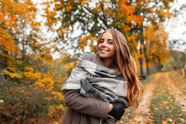 Jeune femme gaie moderne mignonne dans une écharpe tricotée vintage dans une pose chaude manteau d'automne et souriant positivement sur un fond d'arbres au feuillage d'orange. Joyeuse fille modèle promenades dans le parc . — Photo