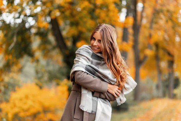 Model van een positieve jonge vrouw in een jas in een gebreide sjaal poserend in een park op een achtergrond van bomen met oranjegeel bladeren.Gelukkig meisje met een leuke glimlach buiten op het platteland in het bos — Stockfoto
