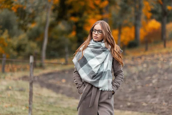 Jeune femme hipster élégante en lunettes dans un manteau à la mode dans une écharpe tricotée dans un champ en dehors de la ville sur un fond d'arbre au feuillage orange. Jolie fille marche à la campagne un jour d'automne . — Photo