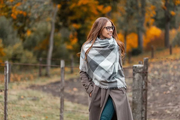 Cheerful young hipster woman in a warm fashionable autumn coat with a knitted scarf in glasses walks through the countryside on the background of a forest with autumn trees. Attractive girl smiles. — Stock Photo, Image