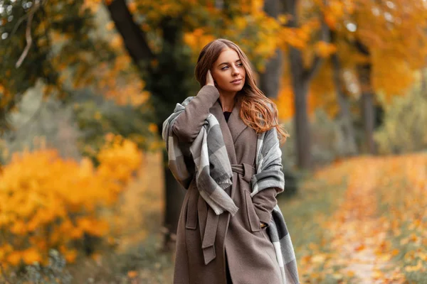 Cute młoda kobieta w ciepłej dzianiny szalik w eleganckim długim płaszczu cieszy się spacer po jesiennym lesie poza miastem. Modny piękny model dziewczyny w stylowej odzieży wierzchniej cieszy się odpoczynkiem w parku. — Zdjęcie stockowe