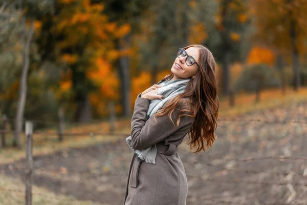 Радостная молодая хипстерша в теплом модном осеннем пальто с вязаным шарфом в очках гуляет по сельской местности на фоне леса с осенними деревьями. Симпатичная милая девушка улыбается. — стоковое фото