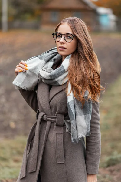 Modelo de una mujer hipster joven con estilo en un abrigo de moda en gafas de moda en una bufanda vintage de punto paseos por el campo. Muchacha linda atractiva. Elegante ropa de mujer de otoño . — Foto de Stock