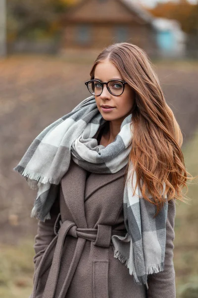 Bonita jovem modelo com óculos em um casaco em um cachecol vintage de malha goza de descanso no campo. Menina muito na moda fica no campo e sorri. Roupas femininas de outono na moda — Fotografia de Stock