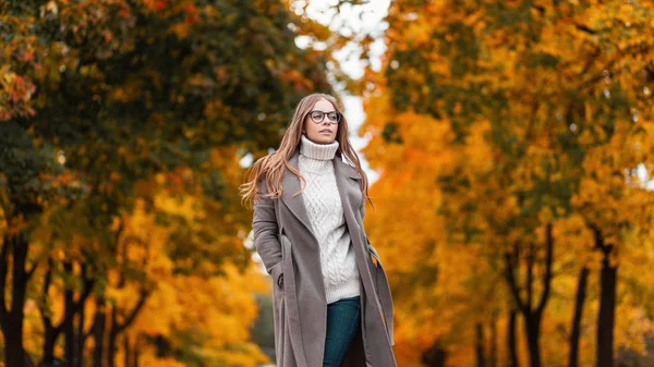 Całkiem stylowa młoda hipsterka w modnym swetrze z dzianiny w eleganckim długim płaszczu w dżinsach w okularach spaceruje po jesiennym parku wśród drzew z pomarańczowymi liśćmi. Piękna dziewczyna na zewnątrz. — Zdjęcie stockowe