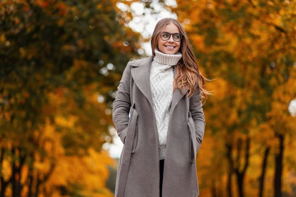 Young cheerful hipster woman in elegant long coat with glasses in a knitted white sweater stands in the park on a warm autumn day. Happy girl enjoys relaxing in the forest. Seasonal women's outerwear. — Stock Photo, Image