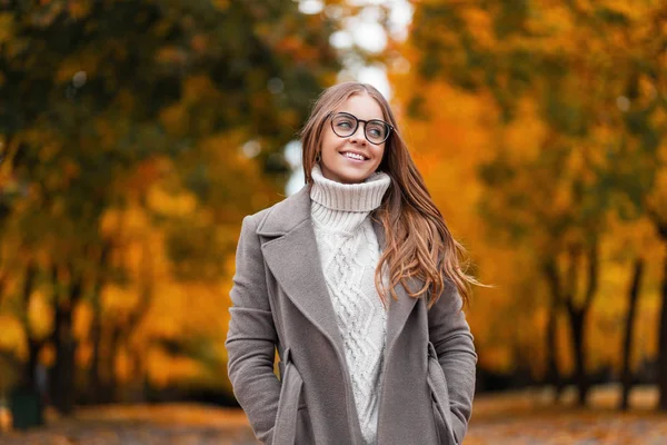 Die süße, fröhliche junge Frau mit dem schönen Lächeln in der Brille im Strickpullover im Herbstmantel entspannt draußen im Wald vor einem Hintergrund von Bäumen mit orange-gelben Blättern. fröhliches Hipster-Mädchen. — Stockfoto