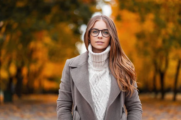 Осенний портрет привлекательной молодой женщины в стильных очках в трикотажном модном белом свитере в элегантном пальто в парке на фоне деревьев с апельсиновыми листьями. — стоковое фото