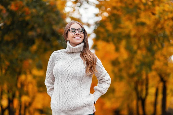 O modelo de uma jovem europeia feliz em um suéter branco na moda tricotado em óculos elegantes com cabelo marrom com um belo sorriso relaxa no parque em um dia de outono. Menina hipster positivo . — Fotografia de Stock