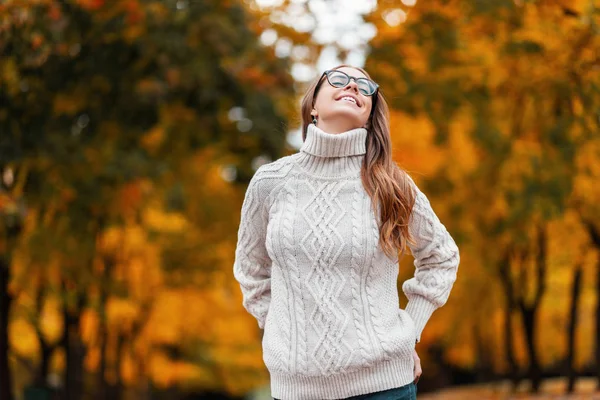 Linda mujer joven divertida con una hermosa sonrisa en gafas en un suéter de punto con un peinado está posando al aire libre en el bosque sobre un fondo de árboles con hojas de color amarillo anaranjado. Chica hipster feliz . — Foto de Stock