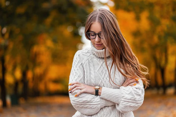 Modelo de una joven europea feliz en un suéter blanco de punto de moda en gafas con estilo con el pelo largo y castaño con se relaja en el parque en un día cálido. Chica hipster de moda. Estilo otoño . — Foto de Stock