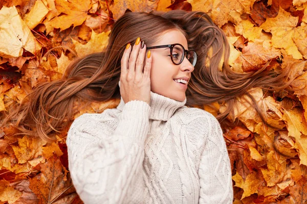 Jovem mulher positiva com um belo sorriso em óculos elegantes em uma camisola de malha branca na moda está descansando na folhagem laranja do outono na floresta. Menina alegre gosta de relaxar ao ar livre em um parque . — Fotografia de Stock