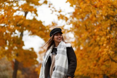 Şık kahverengi ceketli şık şapkalı, zarif, mutlu bir genç kadın. Parkta altın yapraklı ağaçların arka planında poz veren sevimli bir atkısı var. Komik kız dışarıda.