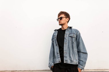 Şık güneş gözlüklü, siyah tişörtlü, klasik kot ceketli modern kentsel adam modeli parlak bir bahar gününde şehirde bir duvarın yanında duruyor. Hipster Amerikalı adam dışarıda. Modaya uygun erkek giyim..